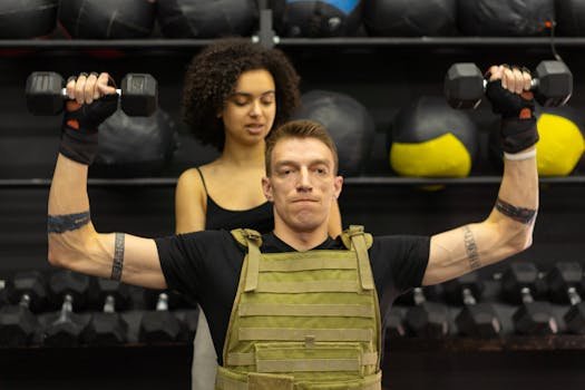 A man in adaptive training gear lifts dumbbells with support from a trainer in a gym.